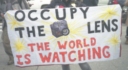Photograph of an individual carrying an "Occupy the Lens" sign